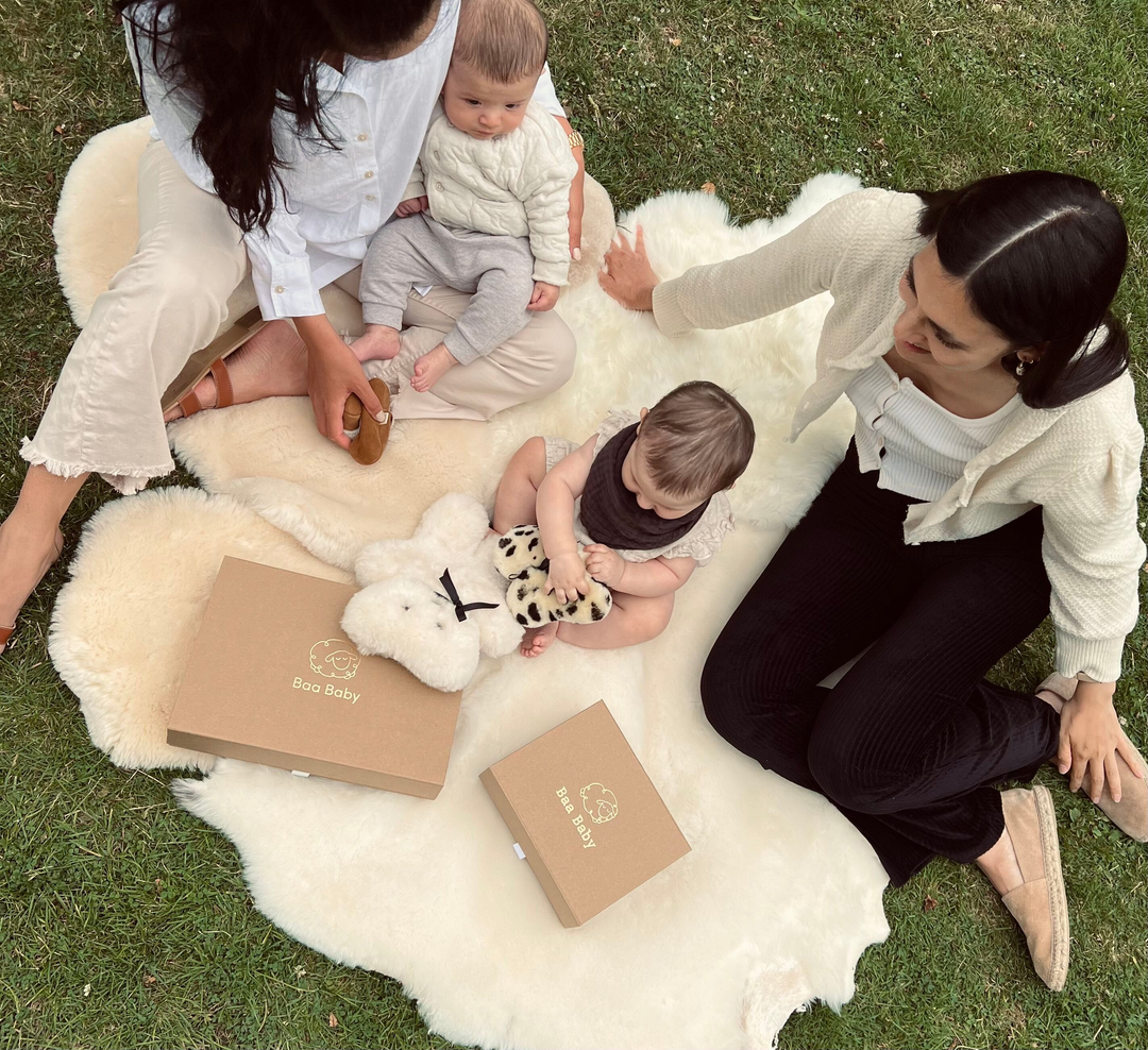 Mum and baby sitting outside on a sheepskin rug sharing baby shower gift boxes with their baby boys including a milk flatout sheepskin teddy bear and natural lambskin booties