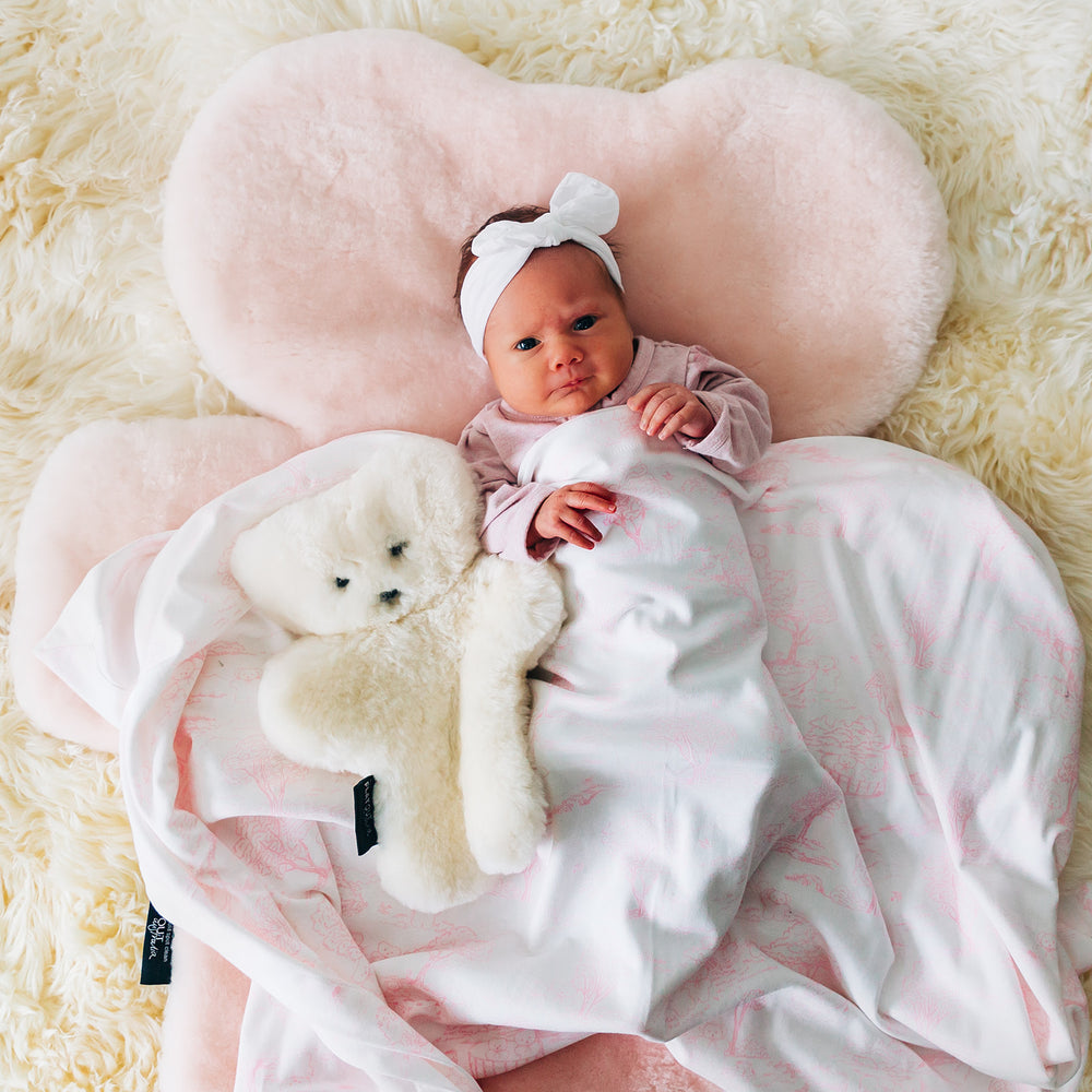 Newborn baby lying on FLATOUT sheepskin bear rug in rosie light pink shade, with a milk sheepskin teddy bear