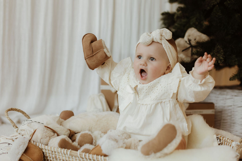 Baby sitting on a sheepskin pram liner in a bassinet wearing teddy merino boucle booties and smiling whilst holding sheepskin lined velcro tabbed booties