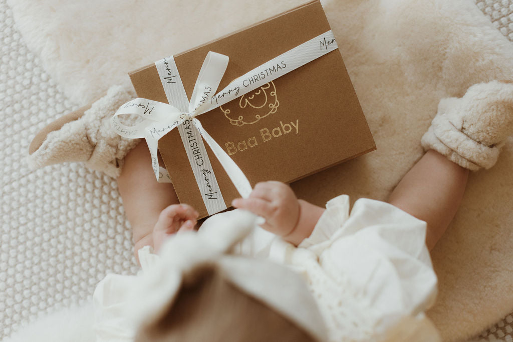 Gift ideas for baby at Christmas showing a toddler opening a luxury gift box with Merry Christmas ribbon containing natural merino boucle booties