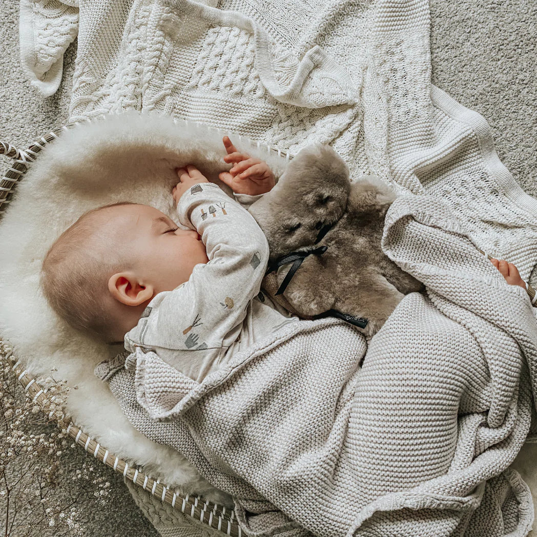 Latte sheepskin FLATOUTbear baby comforter with newborn baby in a seagrass basket snuggled into a milk sheepskin pram liner