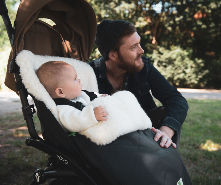 Babyzen Yoyo pram with Fellhof classic sheepskin pram footmuff and baby. Dad sits next to pram and they watch in the park.  