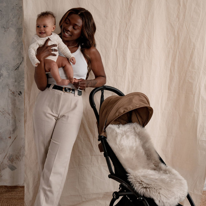 Mum holding baby and smiling, stood next to a yo-yo pram with a neutral brown sheepskin pram liner