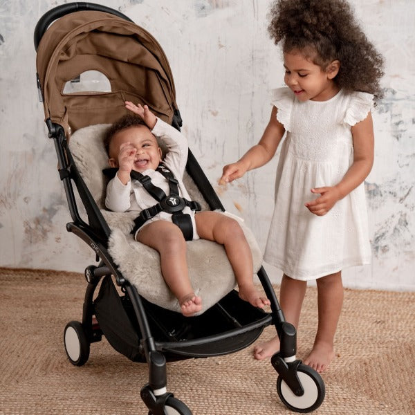 Baby in a pram lined with a neutral sheepskin liner, smiling with his sister stood by the side