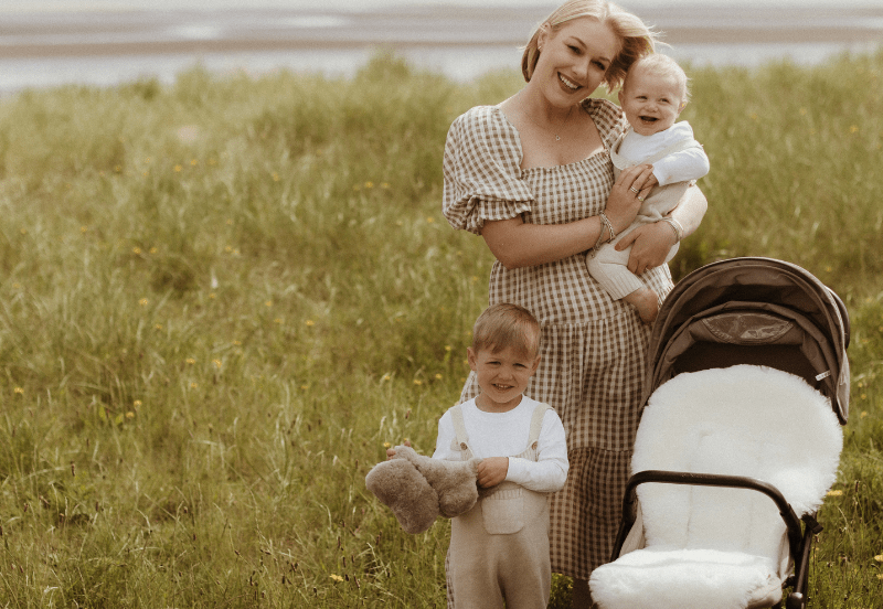 Woman holding a baby and standing next to a child with a teddy bear in a grassy field with a stroller.