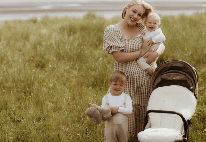Woman holding a baby and standing next to a child with a teddy bear in a grassy field with a stroller.